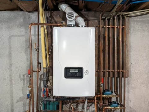 Wall-mounted white tankless water heater with digital display surrounded by copper piping and electrical connections in basement utility room